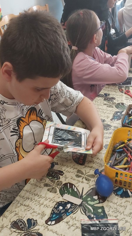 Dzieci uczestniczące w zajęciach plastycznych. Jedno z dzieci wycina papier za pomocą nożyczek, a obok niego siedzi kolejne dziecko. Stół, przy którym pracują, przykryty jest wzorzystym obrusem i pełen materiałów do prac ręcznych – widać kredki, plastikowe woreczki oraz inne przybory. Scena ukazuje twórczą atmosferę zajęć organizowanych w bibliotece.