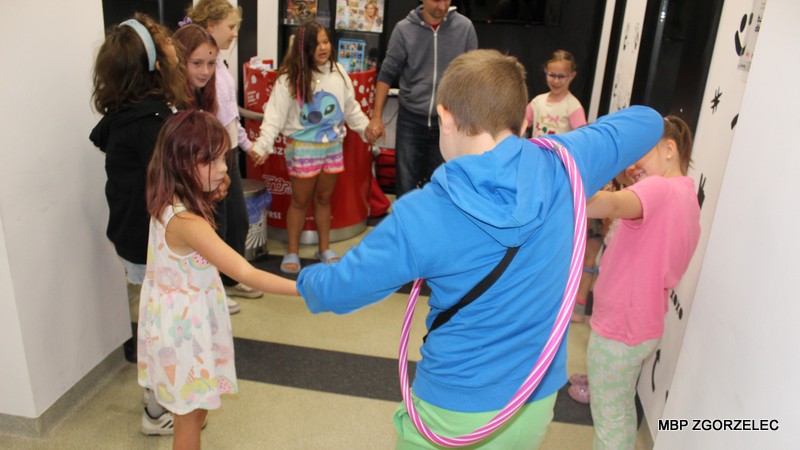 Dzieci i dorosły stoją w kręgu, biorąc udział w zabawie z hula-hop. Na pierwszym planie znajduje się dziecko ubrane w niebieską bluzę z kapturem i zielone spodnie, z różową obręczą hula-hop wokół talii. Scena przedstawia wspólną zabawę i integrację podczas zajęć organizowanych w Bibliotece Multimedialnej.