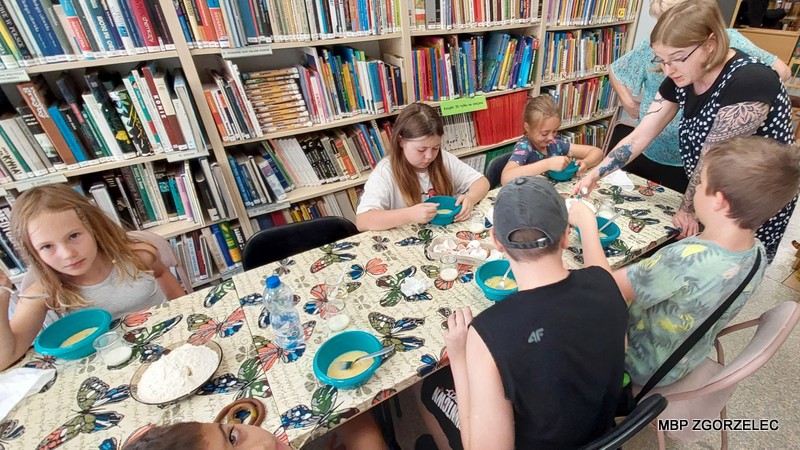 Grupa osób siedzących wokół stołu w przestrzeni bibliotecznej. Stół przykryty jest obrusem z motywem motyli, a na jego powierzchni znajdują się miski i talerze ze składnikami do przygotowania gofrów. W tle widoczne są regały wypełnione książkami, co tworzy przyjazną atmosferę łączącą wspólne spędzanie czasu z otoczeniem literatury.