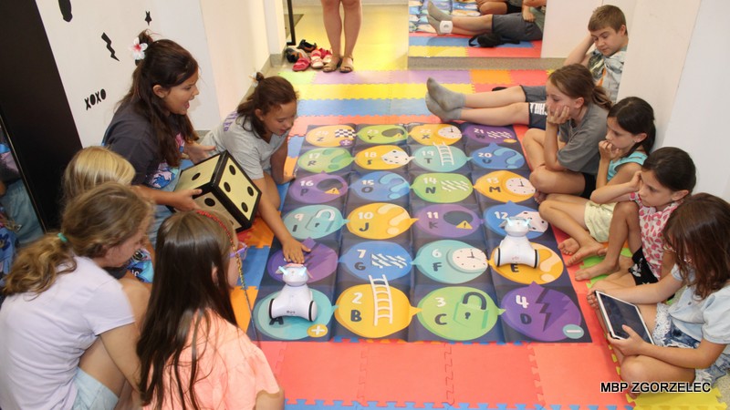 Grupa dzieci siedzących na kolorowych matach piankowych, zgromadzonych wokół dużej planszy do gry edukacyjnej. Plansza zawiera ponumerowane pola od 1 do 20 oraz symbole takie jak drabiny, błyskawice i inne ikony. Na planszy znajdują się dwa małe roboty edukacyjne. Jedno z dzieci trzyma dużą piankową kostkę z czarnymi kropkami, a inne dziecko korzysta z tabletu. Scena odbywa się w bibliotece, ukazując nowoczesne podejście do nauki poprzez zabawę.