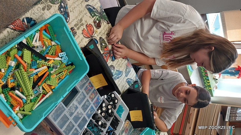 Dwie dziewczynki pracują nad projektem z wykorzystaniem klocków LEGO. Stoją przy stole przykrytym obrusem z motywem motyli, na którym znajdują się pojemniki z różnymi elementami LEGO oraz innymi drobnymi komponentami. Dzieci korzystają z tabletów, które służą im jako źródło instrukcji lub inspiracji do budowy. Scena ukazuje kreatywne i technologiczne podejście do nauki i zabawy w bibliotece.