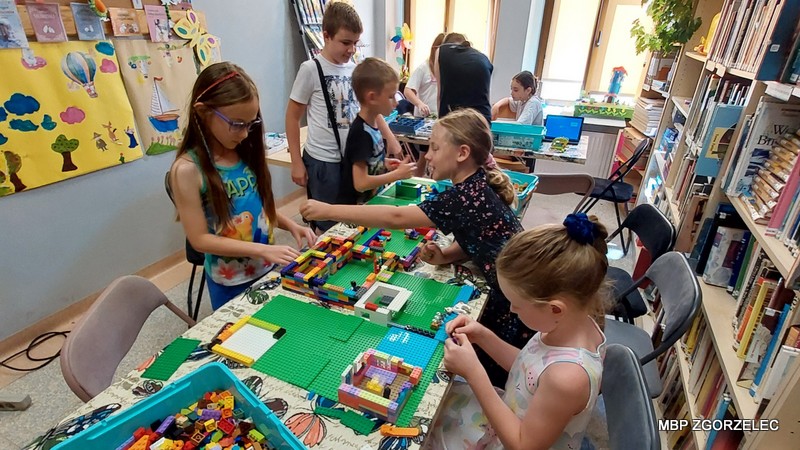 W bibliotece dzieci budują konstrukcje z klocków LEGO, a na stole znajdują się różnorodne elementy oraz częściowo zbudowane modele. W tle widać regały z książkami i materiałami edukacyjnymi, a także kolorowe dekoracje na ścianach, co tworzy inspirujące i kreatywne otoczenie.