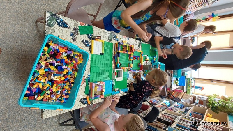 W bibliotece dzieci budują konstrukcje z klocków LEGO, a na stole znajdują się różnorodne elementy oraz częściowo zbudowane modele. W tle widać regały z książkami i materiałami edukacyjnymi, a także kolorowe dekoracje na ścianach, co tworzy inspirujące i kreatywne otoczenie.