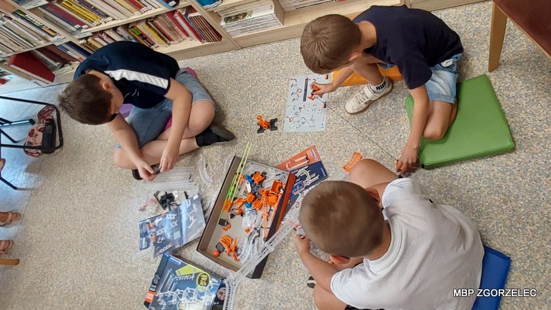 Troje dzieci składa zestaw konstrukcyjny w bibliotece, siedząc na podłodze wśród książek.