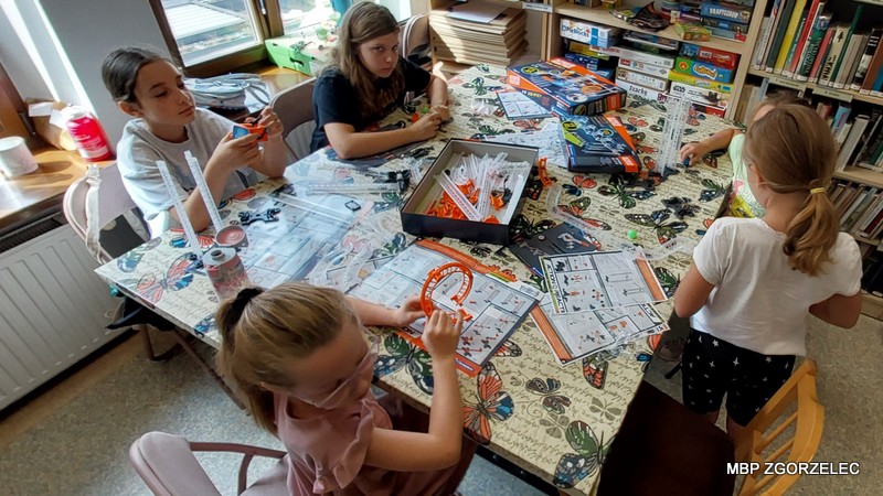 Dzieci budują konstrukcje z plastikowych elementów przy stole w bibliotece, w otoczeniu książek i gier.
