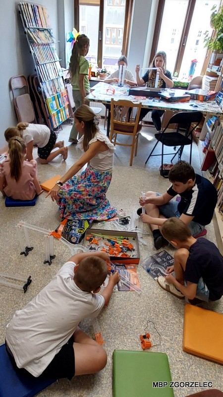 Dzieci i dorosły uczestniczą w zajęciach edukacyjnych w bibliotece polegających na konstruowaniu toru kulkowego, otoczeni są książkami i materiałami dydaktycznymi.