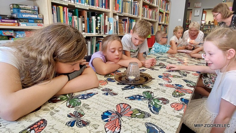 Dzieci uczestniczą w zajęciach - eksperymencie z gasnącą świeczką, przy stole w bibliotece, w tle widoczne książki i gry.