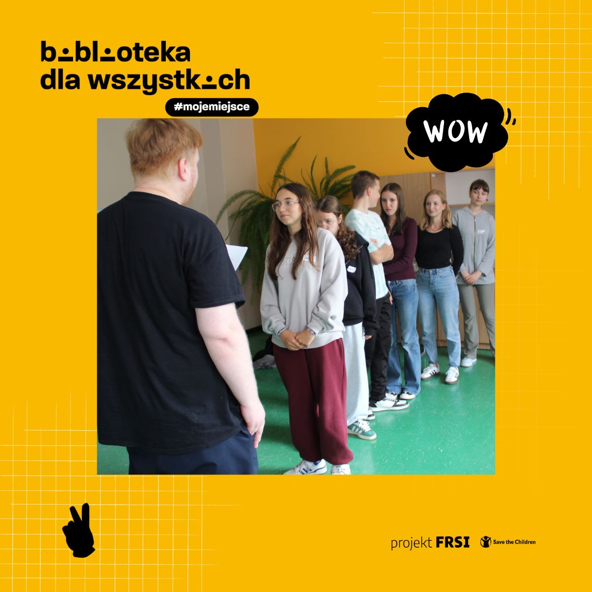 Grupa osób stojących w szeregu w sali lekcyjnej z zieloną podłogą i roślinami w tle. Na zdjęciu widnieją napisy „biblioteka dla wszystkich” oraz hashtag „#mojemiejsce”. W prawym górnym rogu znajduje się grafika z napisem „WOW”, a w dolnym rogu logotypy projektu FRSI i Save the Children.