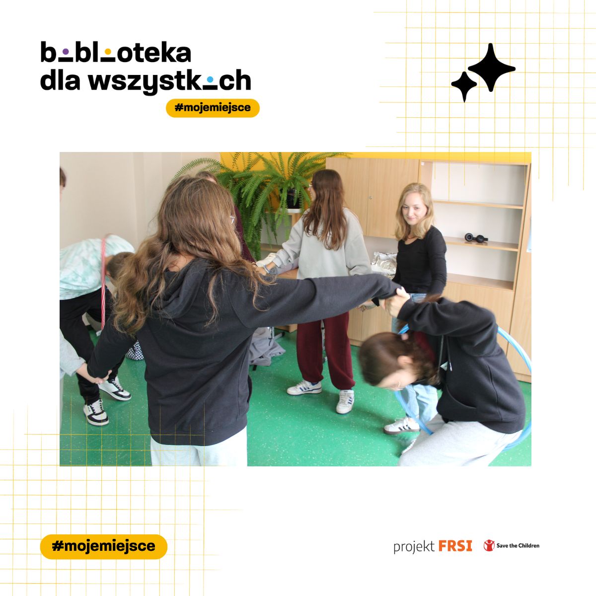 Grupa osób stojących w kręgu i trzymających się za ręce, uczestniczących we wspólnej aktywności z użyciem hula hop. W tle widoczne są rośliny i szafki, bo zajęcia odbywają się w sali lekcyjnej. Na zdjęciu znajdują się napisy: „biblioteka dla wszystkich #mojemiejsce”, „Akademia Kompetencji Przyszłości” oraz logotypy projektu FRSI i Save the Children.