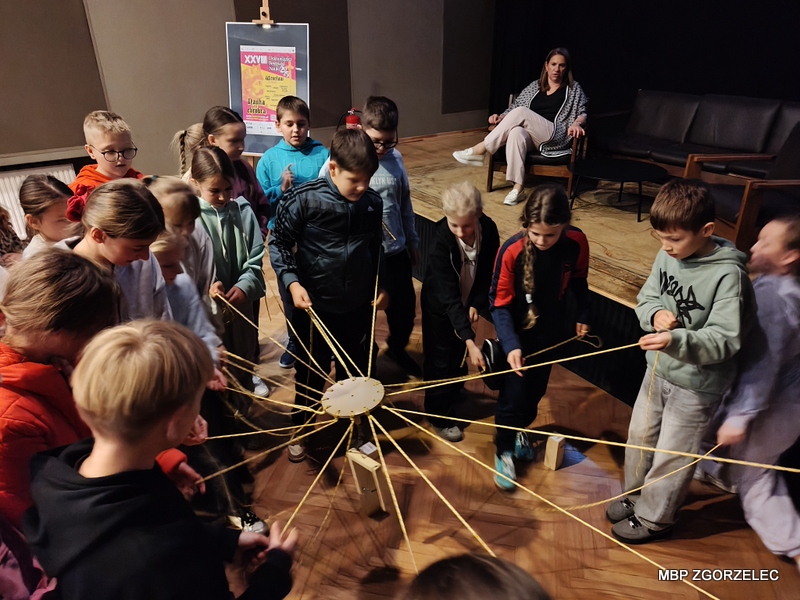 W sali kinowej Miejskiego Domu Kultury w Zgorzelcu dzieci uczestniczą w aktywności zespołowej z wykorzystaniem sznurków i drewnianej konstrukcji. Każde dziecko trzyma sznurek połączony z centralnym elementem, co wskazuje na ćwiczenie współpracy. W sali kinowej Miejskiego Domu Kultury w Zgorzelcu dzieci uczestniczą w aktywności zespołowej z wykorzystaniem sznurków i drewnianej konstrukcji. Każde dziecko trzyma sznurek połączony z centralnym elementem, co wskazuje na ćwiczenie współpracy.