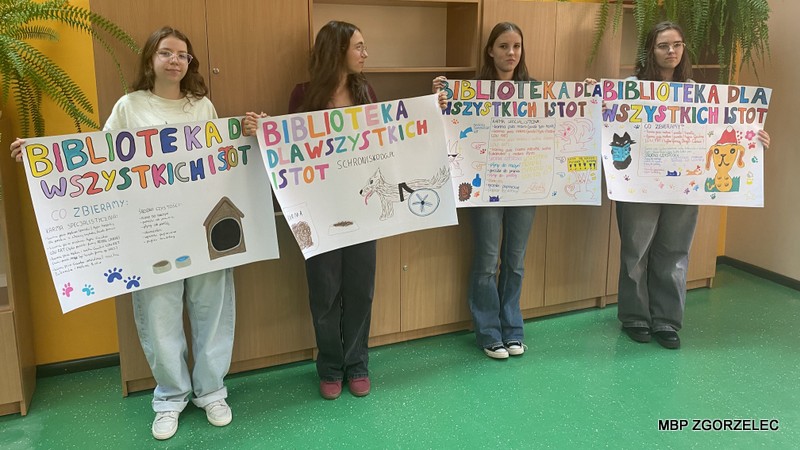 Cztery osoby stojące w sali i prezentujące własnoręcznie wykonane plakaty promujące akcję „Biblioteka dla wszystkich istot”, z kolorowymi napisami i rysunkami zwierząt.