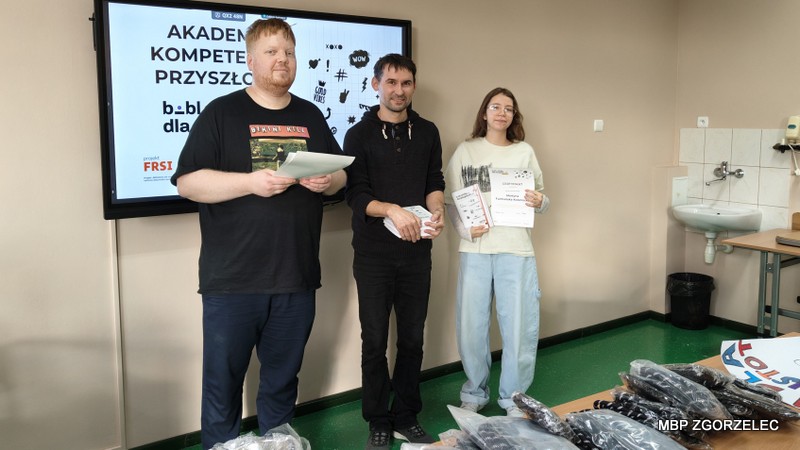 Trzy osoby stojące w sali lekcyjnej przy stole z materiałami na rzecz schroniska. Jedna osoba trzyma notatniki, druga trzyma certyfikaty w ramach programu „Akademia Kompetencji Przyszłości” realizowanego przez bibliotekę i przekazuje je absolwentce programu.