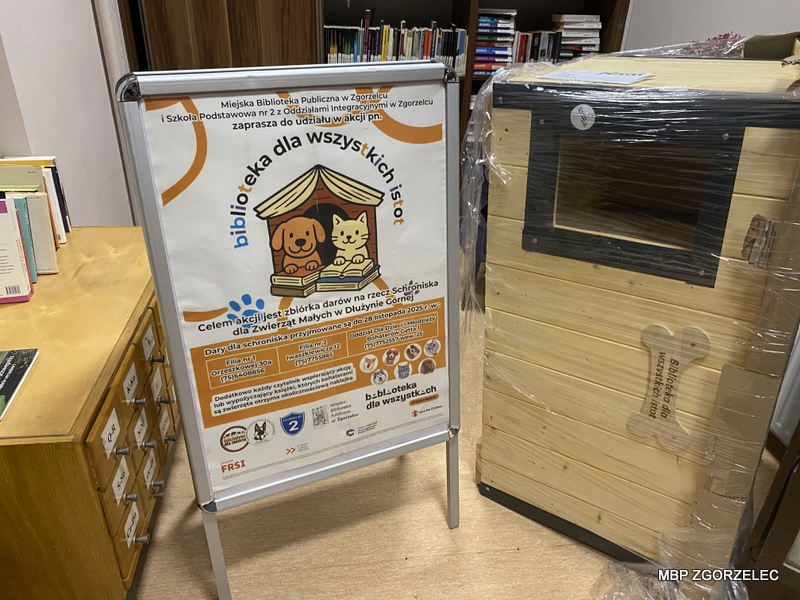 Metalowy stojak z plakatem akcji „Biblioteka dla wszystkich istot” obok dużej drewnianej budy dla psa, owiniętej folią ochronną, przygotowanej do przekazania w ramach zbiórki.
