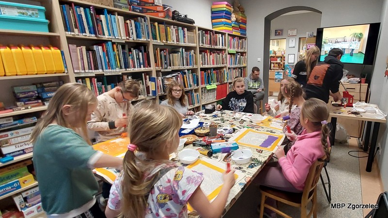 Zdjęcie przedstawia dzieci i&nbsp;młodzież pracujące przy dużym stole w&nbsp;bibliotece, podczas warsztatów plastycznych polegających na malowaniu toreb oraz tworzeniu magnesów i&nbsp;przypinek z&nbsp;motywami zwierząt.