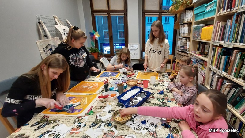 Zdjęcie przedstawia dzieci i&nbsp;młodzież pracujące przy dużym stole w&nbsp;bibliotece, podczas warsztatów plastycznych polegających na malowaniu toreb oraz tworzeniu magnesów i&nbsp;przypinek z&nbsp;motywami zwierząt.