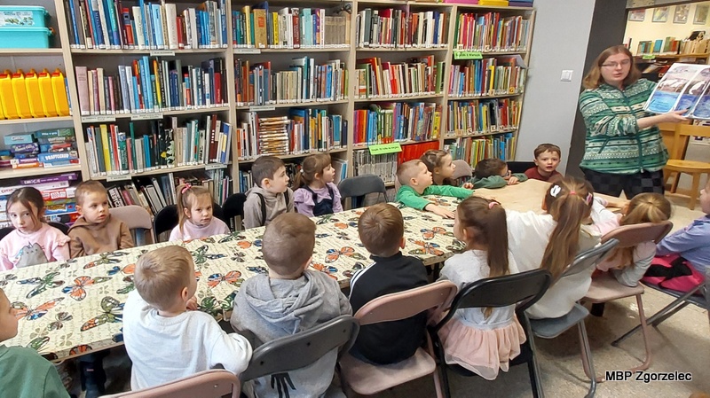 Przedszkolaki siedzą przy długim stole w&nbsp;bibliotece i&nbsp;słuchają opowieści czytanej przez bibliotekarkę, która prezentuje im ilustracje z&nbsp;książki. W&nbsp;tle widoczne są regały pełne książek, a&nbsp;dzieci z&nbsp;uwagą śledzą prezentowaną historię podczas zajęć literackich o&nbsp;tematyce zimowej.