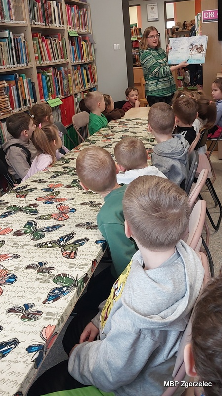 Przedszkolaki siedzą przy długim stole w&nbsp;bibliotece i&nbsp;słuchają opowieści czytanej przez bibliotekarkę, która prezentuje im ilustracje z&nbsp;książki. W&nbsp;tle widoczne są regały pełne książek, a&nbsp;dzieci z&nbsp;uwagą śledzą prezentowaną historię podczas zajęć literackich o&nbsp;tematyce zimowej.