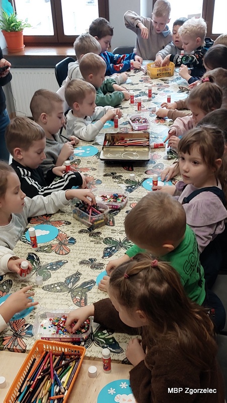 Grupa przedszkolaków bierze udział w&nbsp;zimowych zajęciach literacko‑plastycznych w&nbsp;Oddziale dla Dzieci i&nbsp;Młodzieży MBP w&nbsp;Zgorzelcu. Dzieci siedzą przy długim stole pokrytym kolorowym obrusem i&nbsp;wykonują prace plastyczne, korzystając z&nbsp;kredek, kleju, kolorowego papieru i&nbsp;błyszczących ozdób. Na stole widać koszyczki z&nbsp;przyborami, a&nbsp;dookoła dzieci skupione na tworzeniu „zimowych bombek”, inspirowanych wysłuchaną wcześniej opowieścią o&nbsp;śniegu.
