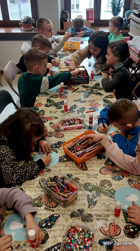 Przy długim stole dzieci wykonują zimowe ozdoby, rysując i&nbsp;dekorując papierowe elementy związane z&nbsp;tematyką zajęć. Przed nimi rozłożone są pudełka z&nbsp;kredkami, pojemniki z&nbsp;błyszczącymi cekinami oraz kleje. Każde dziecko pracuje nad własną pracą, tworząc kolorowe dekoracje. Wokół widać przyjazne otoczenie biblioteczne, a&nbsp;zajęcia odbywają się w&nbsp;atmosferze twórczej pracy.
