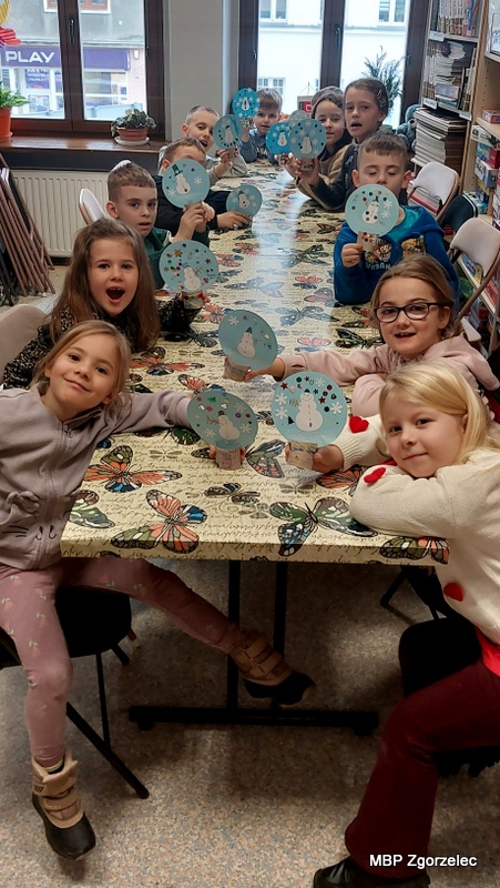 Dzieci siedzą przy stole w&nbsp;bibliotece i&nbsp;trzymają w&nbsp;dłoniach swoje ukończone prace plastyczne – „zimowe bombki” z&nbsp;papieru ozdobione elementami w&nbsp;kształcie płatków śniegu i&nbsp;kolorowymi dekoracjami. Stół jest nakryty motylowym obrusem, a&nbsp;za dziećmi znajdują się duże okna. Dzieci prezentują swoje prace z&nbsp;dumą, podsumowując kreatywną część zajęć zimowych.