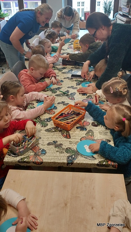 Dzieci siedzą przy stole w&nbsp;bibliotece i&nbsp;ozdabiają papierowe koła za pomocą kredek, kleju i&nbsp;błyszczących elementów. Na środku stołu znajdują się pojemniki z&nbsp;kredkami, cekinami oraz innymi materiałami plastycznymi. Prowadzące osoby z&nbsp;nauczycielami pomagają uczestnikom w&nbsp;pracy i&nbsp;nadzorują przebieg zajęć.