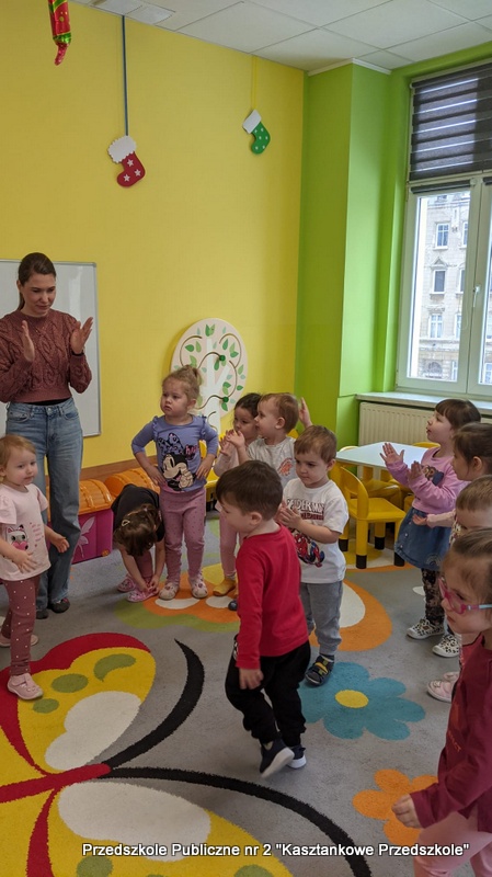 Dzieci stoją i&nbsp;bawią się w&nbsp;kolorowej sali przedszkola. Na podłodze widać dywan w&nbsp;kwiatowe wzory, a&nbsp;w tle znajdują się stoliki, okna i&nbsp;półki z&nbsp;zabawkami. Bibliotekarka stoi przy dzieciach i&nbsp;prowadzi aktywność ruchową, w&nbsp;której uczestniczy cała grupa.