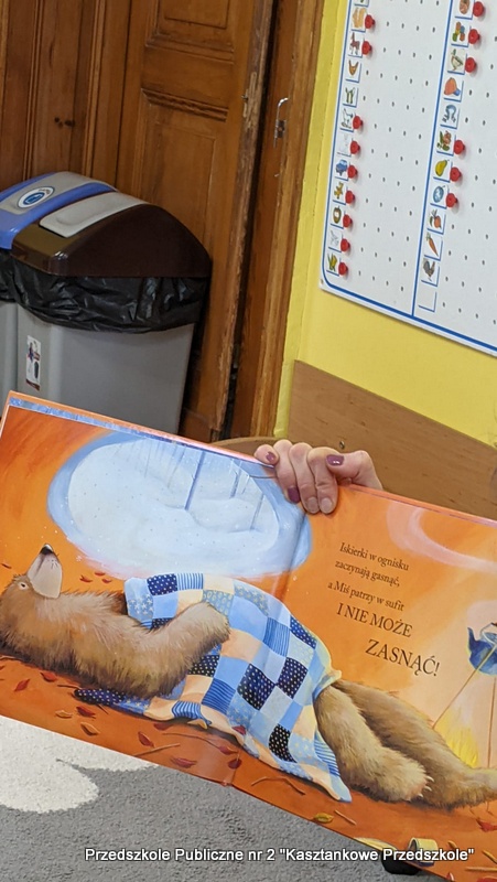 Bibliotekarka trzyma otwartą książkę z&nbsp;ilustracją niedźwiedzia leżącego w&nbsp;łóżku pod niebieską kołdrą. Ilustracja przedstawia zimowy krajobraz z&nbsp;dużym okrągłym oknem i&nbsp;tekstem dotyczącym niedźwiedzia, który nie może zasnąć. W&nbsp;tle widoczna jest żółta ściana, tablica z&nbsp;obrazkami oraz fragment przedszkolnej sali.