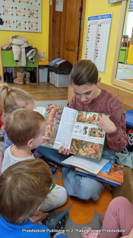 Bibliotekarka siedzi na podłodze w&nbsp;sali przedszkolnej i&nbsp;trzyma otwartą książkę z&nbsp;ilustracjami zwierząt leśnych oraz tekstem. Wokół siedzi kilkoro dzieci, które patrzą na prezentowane strony. W&nbsp;tle widać elementy wyposażenia przedszkola: kolorowe ściany, tablicę, wieszak z&nbsp;ubraniami oraz drzwi. Zdjęcie przedstawia moment wspólnego głośnego czytania w&nbsp;ramach zajęć edukacyjnych.