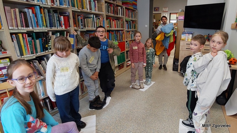 Dzieci uczestniczą w&nbsp;zabawie prowadzonej przez animatora w&nbsp;dziale dziecięcym biblioteki. Stoją na kartkach papieru ułożonych na podłodze, wykonując zadania ruchowe. W&nbsp;tle widać regały z&nbsp;książkami, dekoracje oraz sprzęt dydaktyczny.