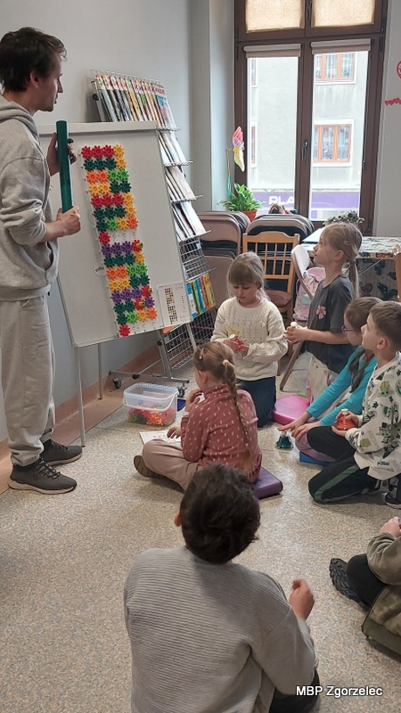 Dzieci siedzą na podłodze i&nbsp;patrzą na prowadzącego, który prezentuje wzór ułożony z&nbsp;kolorowych klocków na tablicy stojącej na sztaludze. Zajęcia odbywają się w&nbsp;sali biblioteki wyposażonej w&nbsp;regały, materiały edukacyjne i&nbsp;stoliki do pracy twórczej.