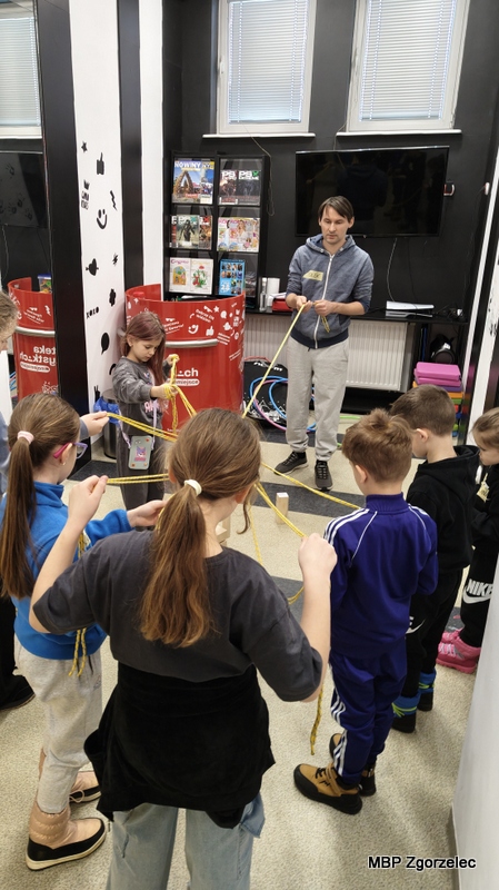 Grupa dzieci stoi w&nbsp;kole, trzymając w&nbsp;rękach długie sznurki używany w&nbsp;zabawie integracyjnej prowadzonej przez animatora. Zajęcia odbywają się w&nbsp;Mediatece Miejskiej Biblioteki Publicznej w&nbsp;Zgorzelcu wyposażonej w&nbsp;regały z&nbsp;magazynami, gry oraz miejsce do aktywności.