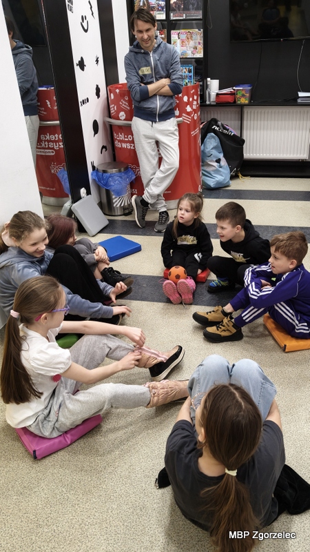 Grupa dzieci siedzi na podłodze w&nbsp;kręgu, rozmawiając i&nbsp;uczestnicząc w&nbsp;spokojnej części zajęć prowadzonych przez animatora.