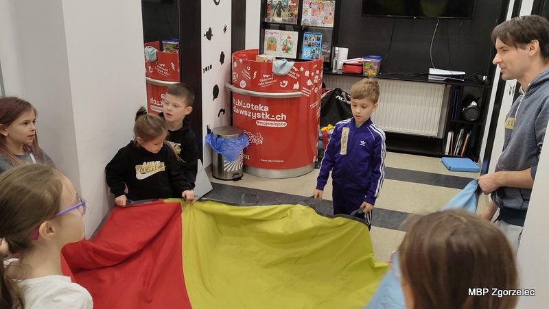 Kilku uczestników zajęć stoi w&nbsp;kole, trzymając dużą, wielokolorową chustę animacyjną. Obok nich stoi prowadzący zajęcia. Sala jest udekorowana przyjaznymi grafikami i&nbsp;elementami przeznaczonymi do działań animacyjnych w&nbsp;bibliotece.