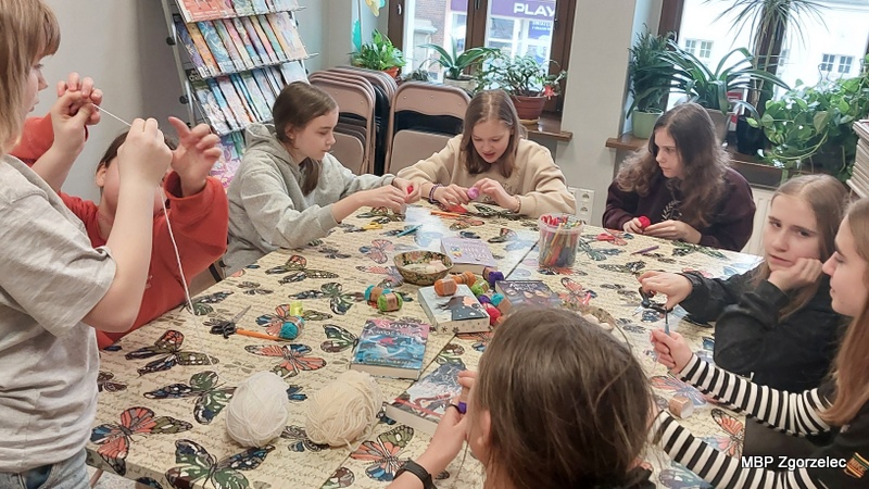 Uczestnicy warsztatów wykonują ozdoby z&nbsp;włóczki przy dużym stole w&nbsp;bibliotece.