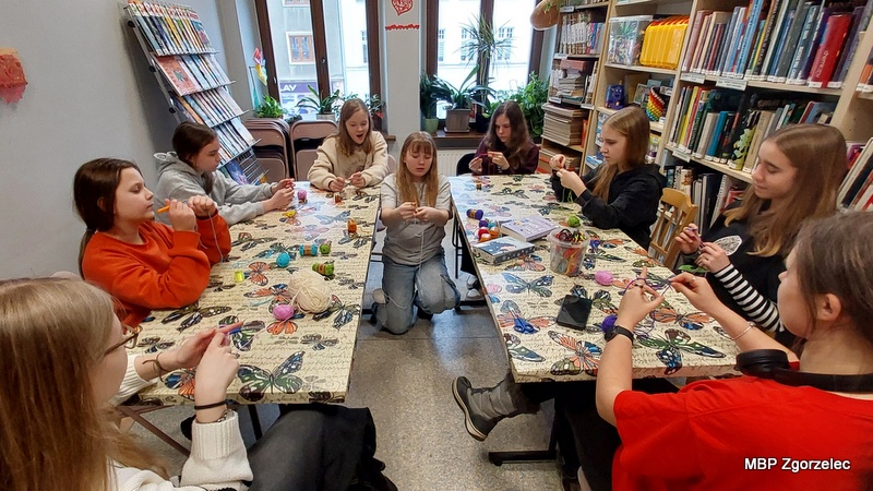 Instruktorka stojąca przy stole pokazuje uczestnikom, jak wykonywać ozdoby z włóczki. Wokół niej młodzież siedzi przy stole z materiałami do prac ręcznych: motkami włóczki, nożyczkami, pisakami oraz gotowymi elementami ozdobnymi. W tle widać regały z książkami działu bibliotecznego.