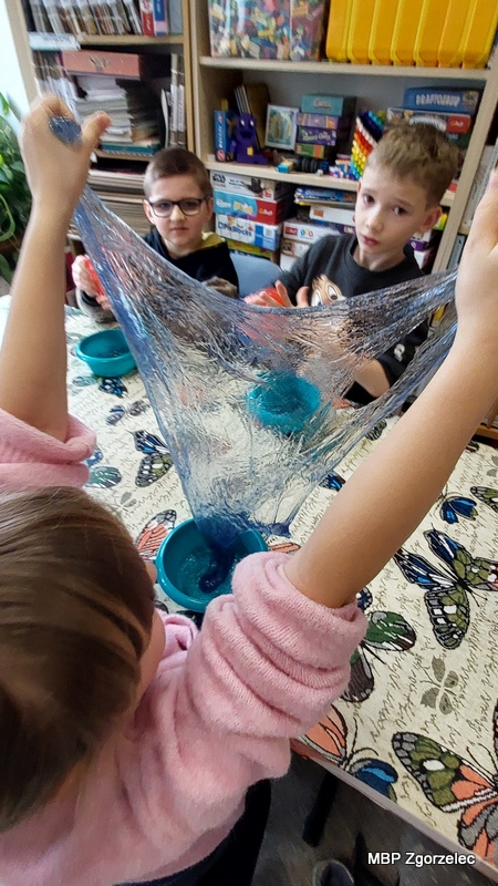 Dziecko trzyma i&nbsp;rozciąga przezroczystą, błyszczącą masę typu slime nad miseczką, tworząc długi, elastyczny fragment masy. Na stole znajdują się kolorowe przybory plastyczne i&nbsp;pojemniki, a&nbsp;w tle widać regały z&nbsp;książkami oraz gry edukacyjne.