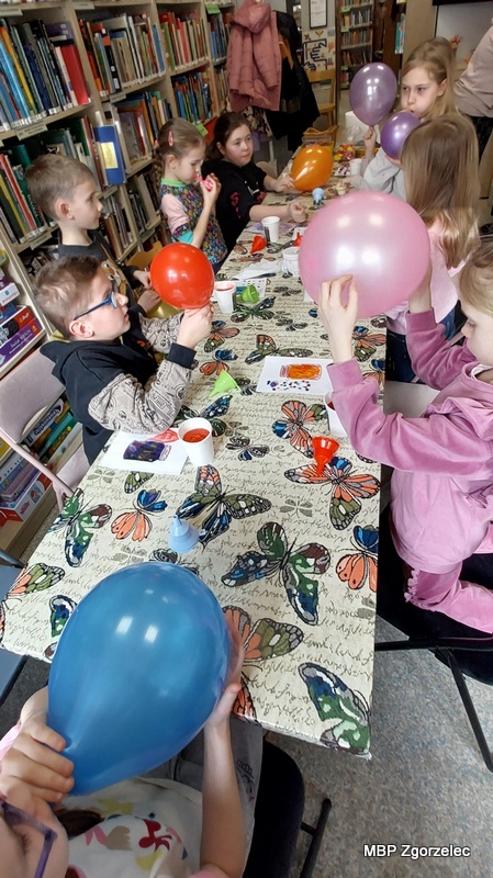Dzieci siedzą przy stole i&nbsp;nadmuchują kolorowe balony w&nbsp;ramach zabawy lub eksperymentu prowadzonego podczas zajęć. Na stole leżą narzędzia plastyczne oraz przygotowane kartki. Po obu stronach stołu znajdują się regały z&nbsp;książkami działu dziecięcego.