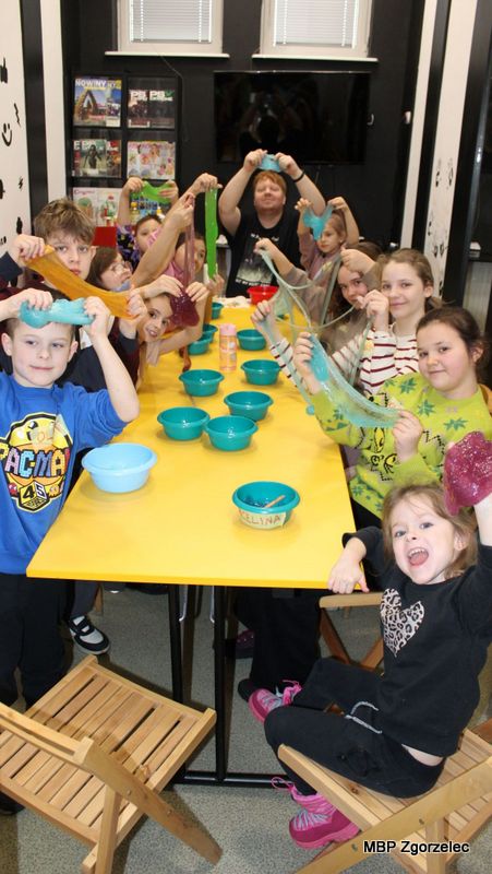 Dzieci siedzą i&nbsp;stoją wokół długiego stołu, trzymając wysoko rozciągniętą masę slime w&nbsp;różnych kolorach. Przed nimi znajdują się miseczki i&nbsp;materiały używane podczas warsztatów kreatywno-sensorycznych. Zajęcia mają formę wspólnej zabawy i&nbsp;eksperymentowania.