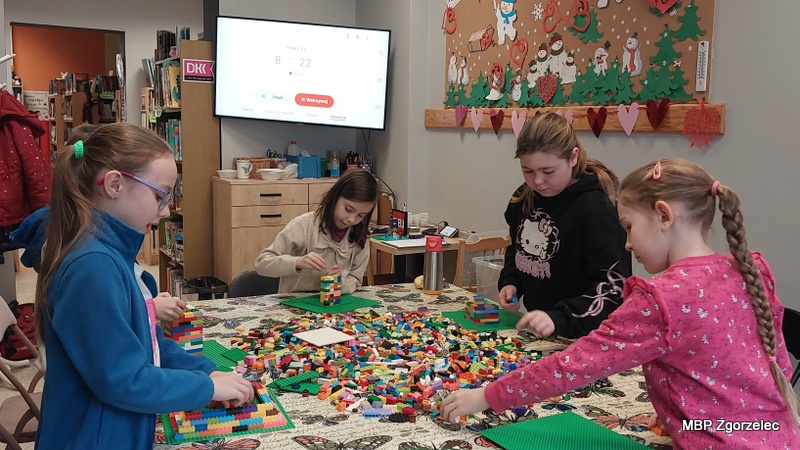 Kilka dzieci siedzi przy stole pokrytym obrusem w&nbsp;motyle i&nbsp;tworzy budowle z&nbsp;kolorowych klocków LEGO. Na środku stołu leży duża sterta klocków, a&nbsp;uczestnicy korzystają z&nbsp;zielonych płytek jako podstaw. Zajęcia odbywają się w&nbsp;sali bibliotecznej z&nbsp;książkami i&nbsp;dekoracjami na ścianach.