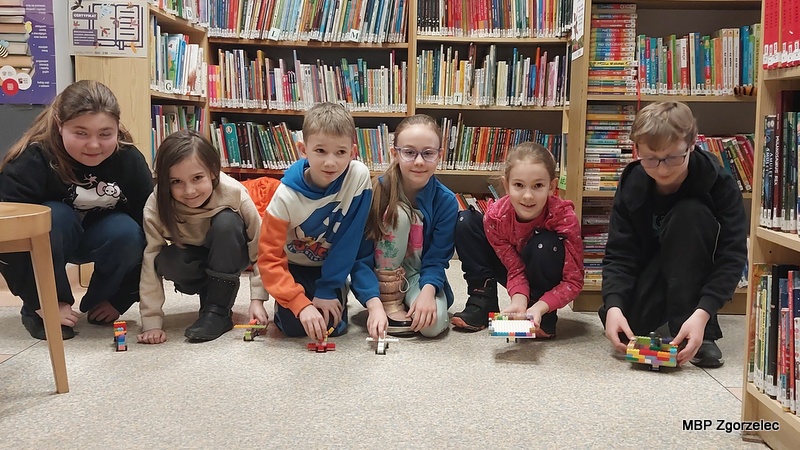 Dzieci klęczą na podłodze między regałami bibliotecznymi i&nbsp;ustawiają przed sobą własnoręcznie zbudowane pojazdy z&nbsp;klocków LEGO. W&nbsp;tle widać półki pełne książek oraz elementy dekoracyjne charakterystyczne dla przestrzeni dziecięcej biblioteki.