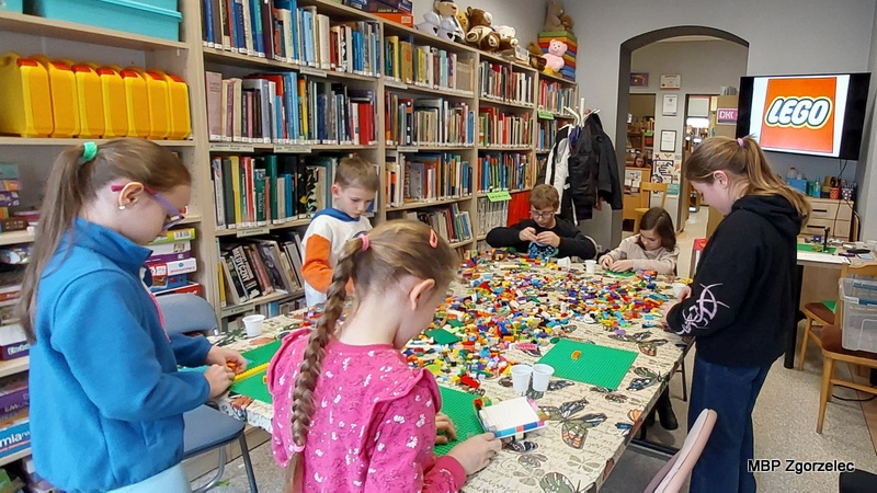 Dzieci stoją i&nbsp;siedzą wokół dużego stołu, na którym znajdują się setki klocków LEGO. Każde z&nbsp;nich pracuje nad swoją konstrukcją, korzystając z&nbsp;płytek i&nbsp;kolorowych elementów. Tło stanowią wysokie regały z&nbsp;książkami oraz ekran z&nbsp;logo LEGO.
