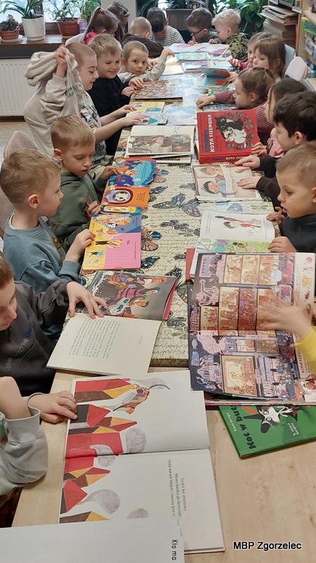 Dzieci siedzą po obu stronach długiego stołu w&nbsp;bibliotece, przeglądając różnorodne książki dla najmłodszych. Na stole rozłożono liczne kolorowe publikacje: książki obrazkowe, opowiadania oraz komiksy. Dzieci otwierają książki i&nbsp;oglądają ilustracje. W&nbsp;tle widać półki z&nbsp;księgozbiorami oraz fragment okna. Zdjęcie wykonano 16 lutego 2026 r. w&nbsp;Oddziale dla Dzieci i&nbsp;Młodzieży MBP Zgorzelec podczas zajęć związanych ze Światowym Dniem Kota.