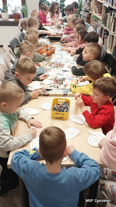Przy długim stole w&nbsp;bibliotece siedzi grupa dzieci wykonujących prace plastyczne. Na stole leżą papierowe szablony kotów, kredki świecowe oraz pudełko z&nbsp;przyborami plastycznymi. Dzieci zajmują się kolorowaniem i&nbsp;dekorowaniem przygotowanych szablonów. W&nbsp;tle widać regały z&nbsp;książkami oraz okno wpuszczające naturalne światło. Zdjęcie wykonano podczas części artystycznej zajęć z&nbsp;okazji Światowego Dnia Kota w&nbsp;Oddziale dla Dzieci i&nbsp;Młodzieży MBP Zgorzelec, 16 lutego 2026 r.