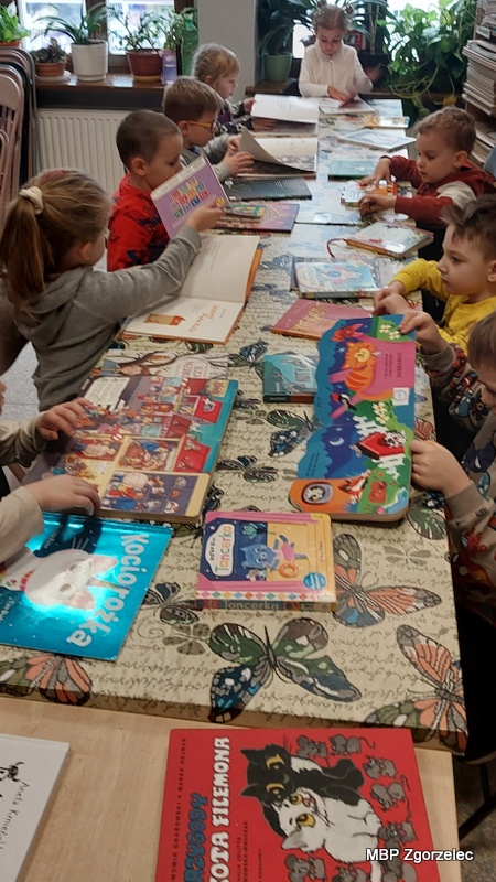 Dzieci siedzą przy długim stole przykrytym obrusem w&nbsp;motyle, a&nbsp;przed nimi leżą liczne książki obrazkowe o&nbsp;kotach i&nbsp;innych bohaterach bajek. Każde dziecko ma przed sobą jedną lub kilka książek i&nbsp;przegląda ilustracje. Na pierwszym planie widać okładki publikacji w&nbsp;żywych kolorach. W&nbsp;tle znajdują się regały z&nbsp;książkami i&nbsp;rośliny doniczkowe na parapecie. Zdjęcie wykonano 17 lutego 2026 r., podczas spotkania w&nbsp;bibliotece.