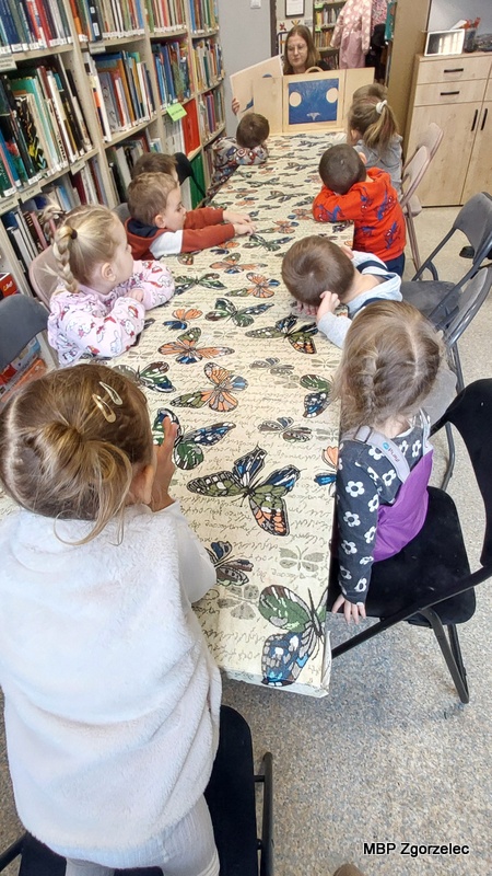 Dzieci siedzą na krzesłach po obu stronach długiego stołu, zwrócone w&nbsp;stronę małej drewnianej sceny kamishibai ustawionej na końcu stołu. Na scenie widoczna jest ilustracja prezentowana podczas opowiadania. Wokół widać regały wypełnione książkami. Obrus pokrywający stół ozdobiony jest wzorem kolorowych motyli. Zdjęcie wykonano 17 lutego 2026 r., jako dokumentację zajęć z&nbsp;prezentacją kamishibai.