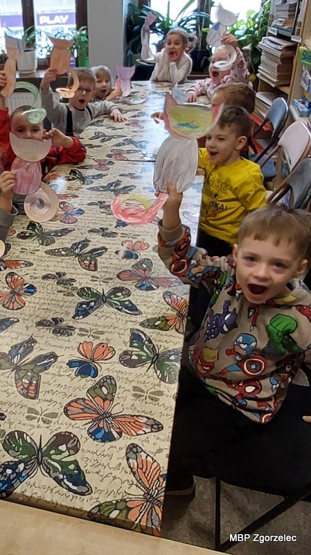 Dzieci siedzą przy stole w&nbsp;bibliotece i&nbsp;trzymają w&nbsp;dłoniach papierowe prace plastyczne w&nbsp;kształcie kotów, które wcześniej pokolorowały. W&nbsp;powietrzu uniesione są różnokolorowe rysunki przedstawiające koty o&nbsp;spiralnych ogonach. Na stole leży obrus z&nbsp;motylami, a&nbsp;obok znajdują się krzesła i&nbsp;regały z&nbsp;książkami. Zdjęcie wykonano 17 lutego 2026 r., podczas zajęć plastycznych związanych ze Światowym Dniem Kota.