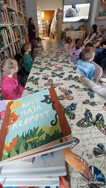 Dzieci siedzą przy długim stole w&nbsp;bibliotece, skierowane w&nbsp;stronę ekranu, na którym wyświetlany jest film edukacyjny o&nbsp;kotach domowych. Na stole leżą liczne książki dla najmłodszych, w&nbsp;tym widoczna na pierwszym planie publikacja „Koty mają kłopoty”. Bibliotekarka stoi przy regale i&nbsp;prowadzi zajęcia. W&nbsp;tle znajdują się wysokie półki z&nbsp;książkami oraz stojące krzesła. Zdjęcie wykonano 23 lutego 2026 r. podczas zajęć z&nbsp;okazji Światowego Dnia Kota.