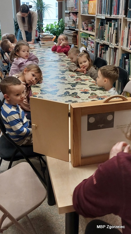 Dzieci siedzą po obu stronach długiego stołu w&nbsp;bibliotece i&nbsp;oglądają przedstawienie kamishibai. Na pierwszym planie znajduje się drewniana skrzynka kamishibai z&nbsp;widoczną ilustracją kota. W&nbsp;tle widać półki wypełnione książkami oraz rośliny ustawione przy oknie. Zdjęcie wykonano 23 lutego 2026 r. w&nbsp;trakcie prezentacji teatrzyku obrazkowego.