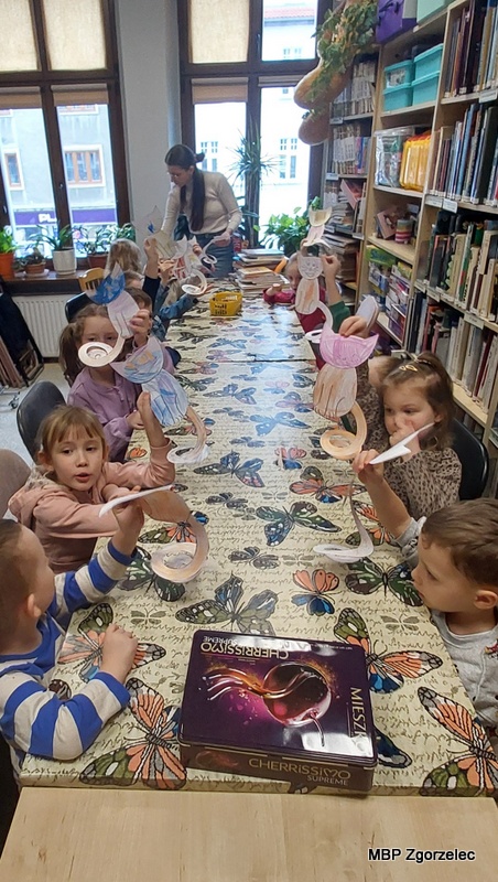 Przy długim stole w&nbsp;bibliotece siedzi grupa dzieci trzymających w&nbsp;dłoniach wykonane przez siebie papierowe kotki o&nbsp;spiralnych ogonach. W&nbsp;tle widoczne są duże okna, rośliny doniczkowe oraz regały z&nbsp;książkami. Na stole leży pudełko z&nbsp;materiałami. Zdjęcie wykonano 23 lutego 2026 r. podczas zajęć plastycznych nawiązujących do Światowego Dnia Kota.