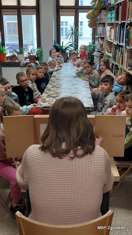 Dzieci siedzą po obu stronach długiego stołu w&nbsp;czytelni biblioteki, zwrócone w&nbsp;stronę prowadzącej, która siedzi przy małej drewnianej skrzynce kamishibai i&nbsp;prezentuje ilustracje. Stół nakryty jest obrusem we wzór motyli. W&nbsp;tle widoczne są wysokie regały wypełnione książkami oraz duże okna z&nbsp;widokiem na zabudowania po drugiej stronie ulicy. Zdjęcie wykonano 24 lutego 2026 r. podczas prezentacji teatrzyku kamishibai w&nbsp;Oddziale dla Dzieci i&nbsp;Młodzieży.